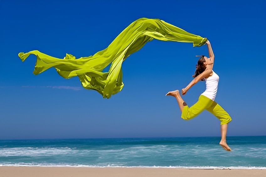 Frau springt energiegeladen am Strand mit wehendem grünen Tuch - Symbol für Vitalität Freiheit und Lebensfreude bei strahlend blauem Himmel